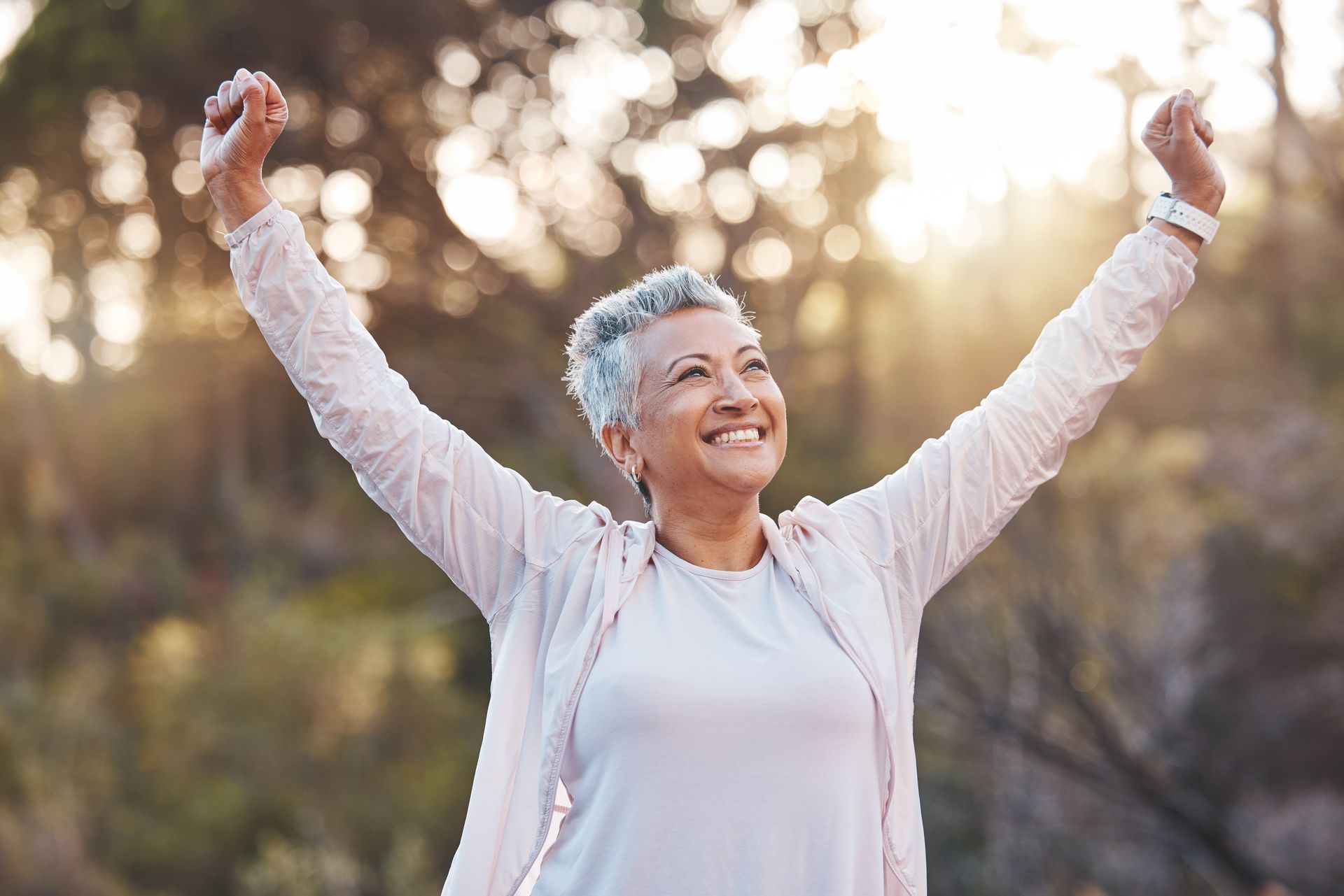 Smiling person with arms raised outdoors in soft sunlight, wearing a white jacket