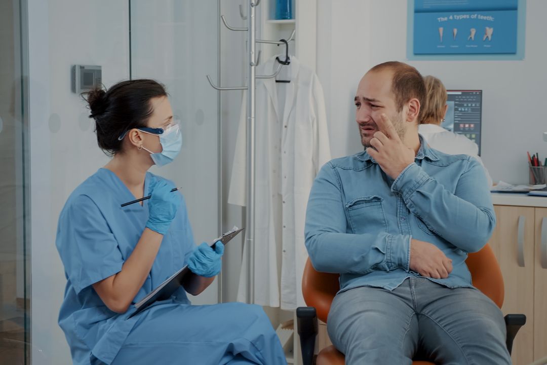 Nurse in blue scrubs talks with a seated patient in a clinic room, both wearing face masks.