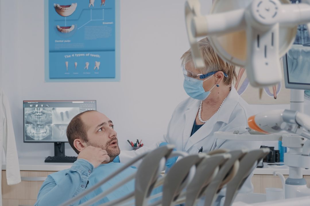 Dentist consulting seated patient in a bright dental clinic, with X-ray monitor and treatment equipment nearby