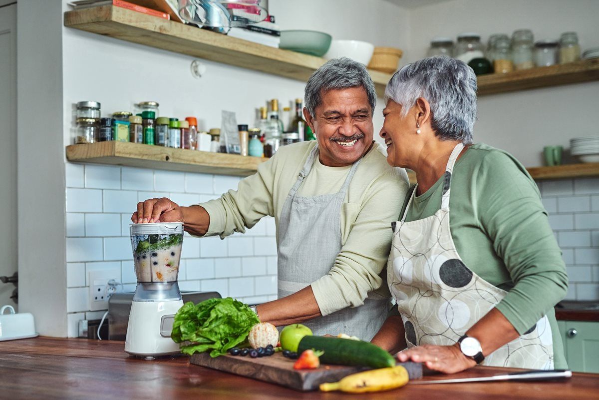 Smiling couple making a smoothie in a kitchen. Vegetables and fruit on counter.
