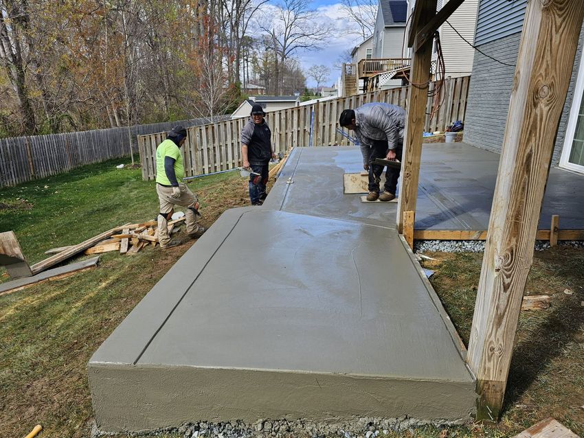 Three people work on a newly poured concrete patio and step in a residential backyard on a sunny day.