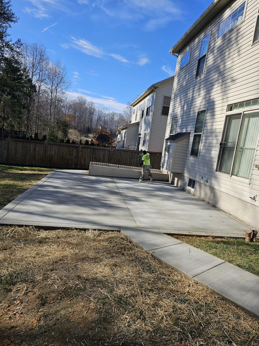 A worker stands on a freshly poured concrete patio in the backyard of a beige, multi-story house.