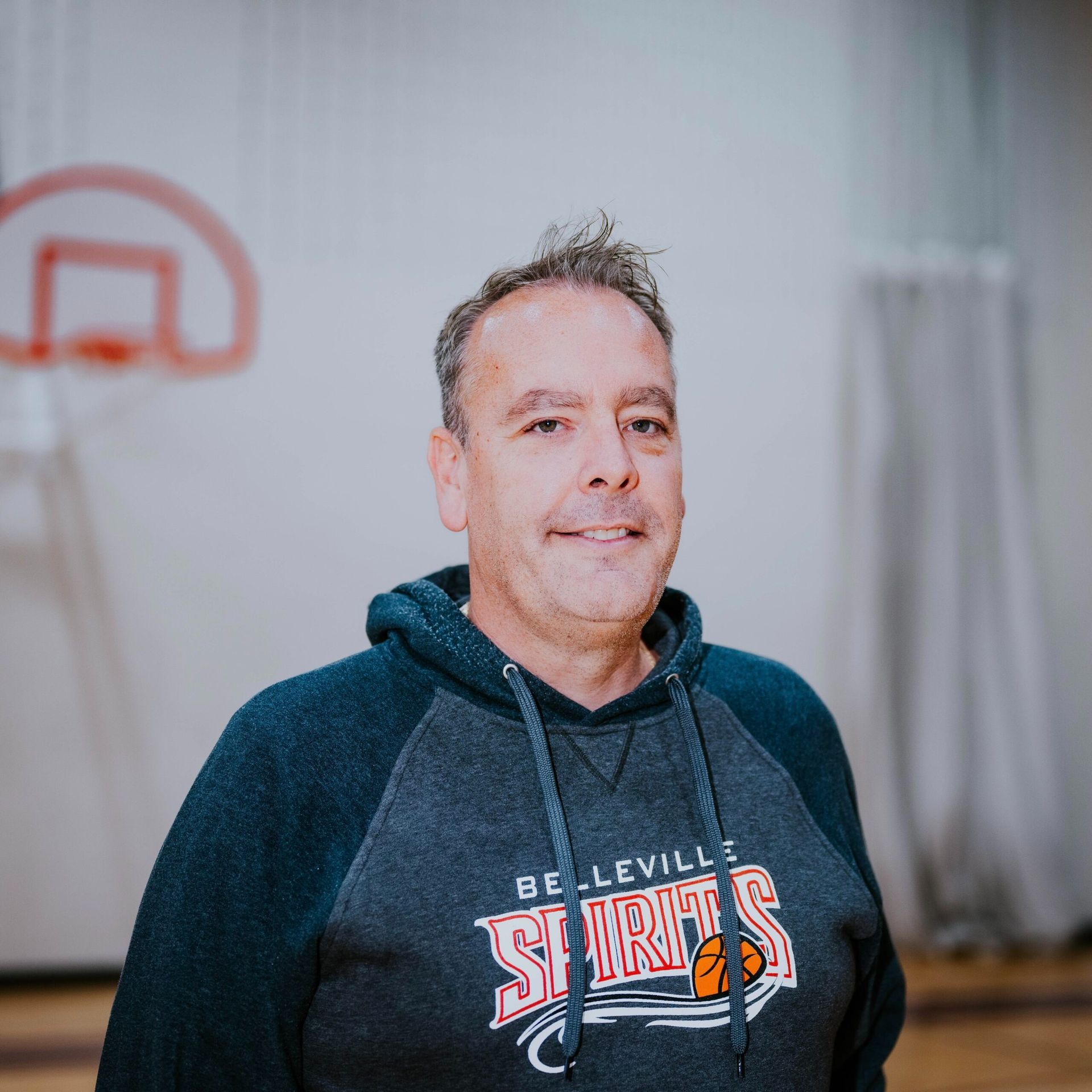 Man in a dark hoodie standing indoors, with a basketball hoop blurred in the background