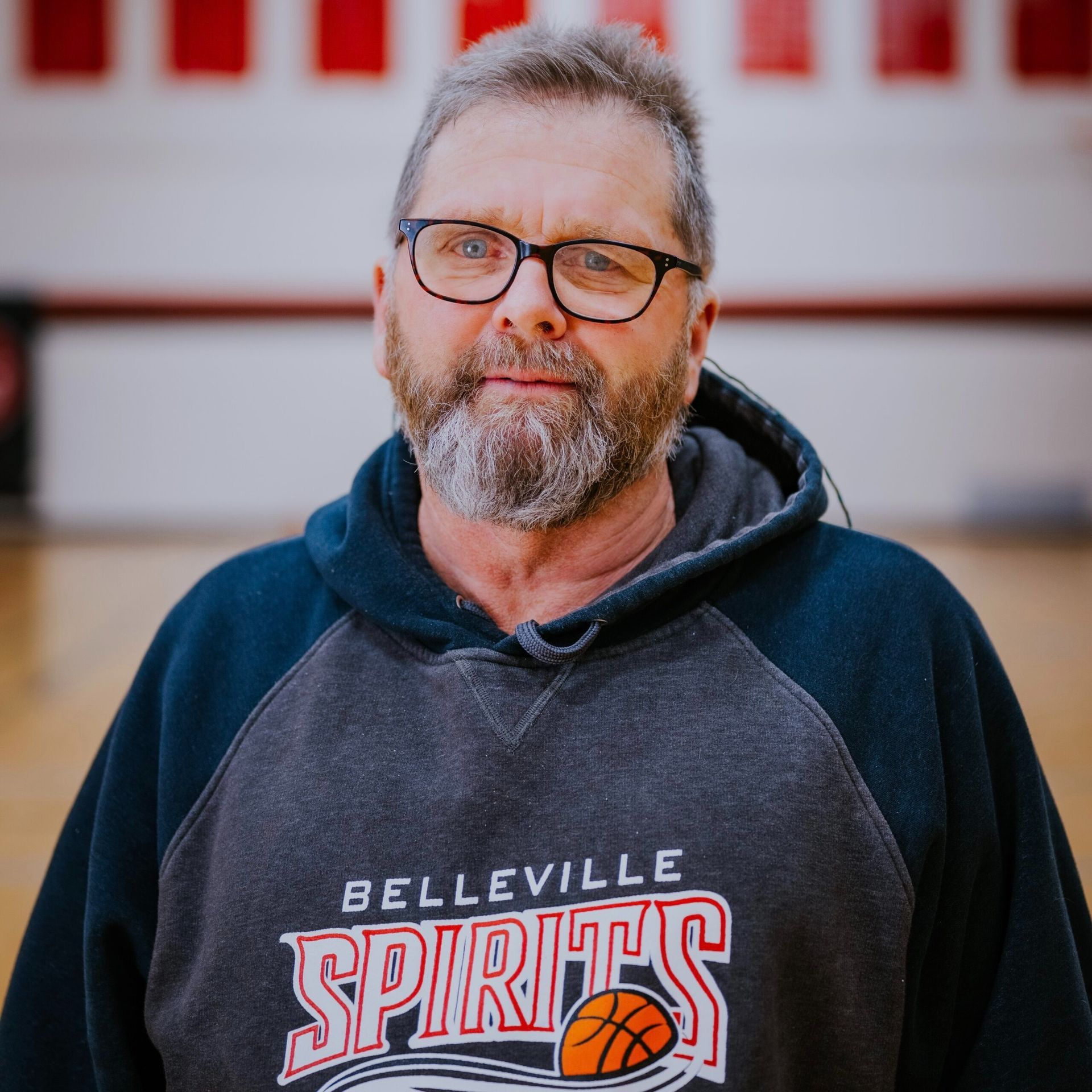 Man wearing glasses and a Belleville Spirits hoodie, standing outdoors with a blurred red-and-white background