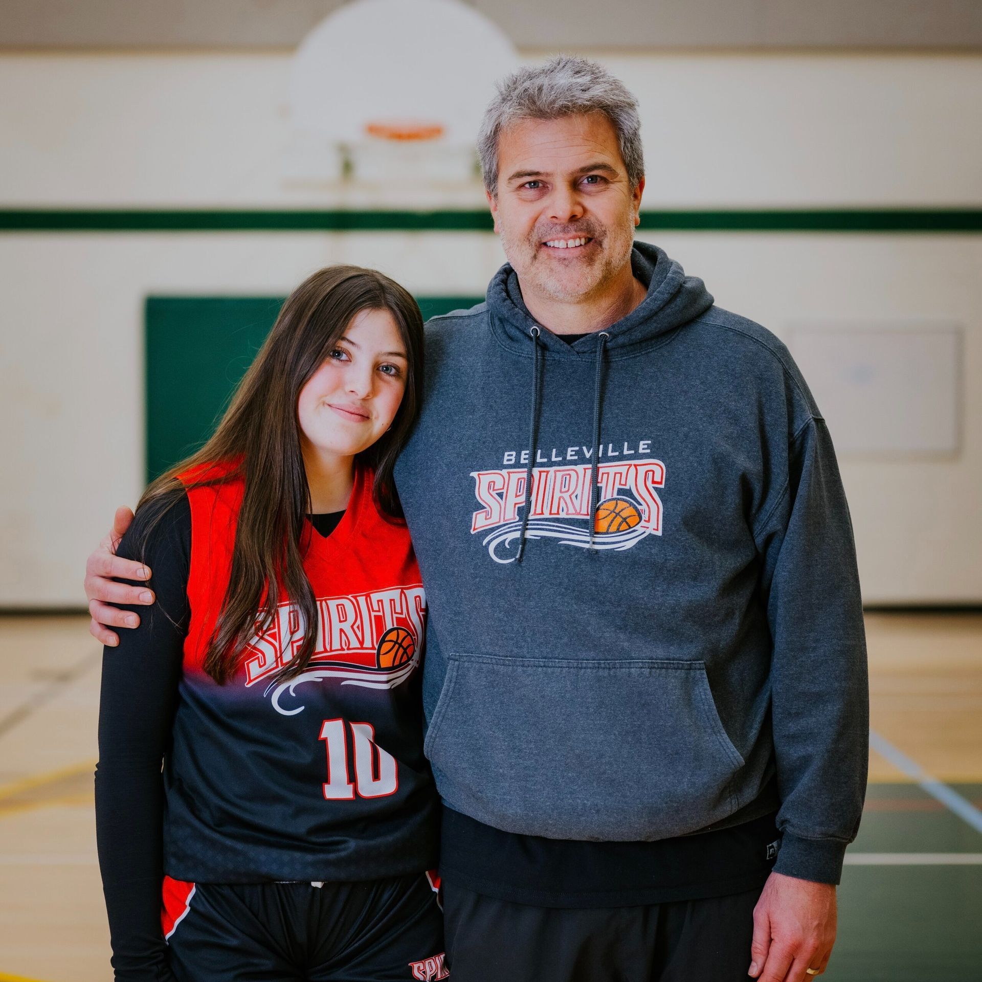 Two people in sports hoodies stand together in a gym, smiling at the camera.