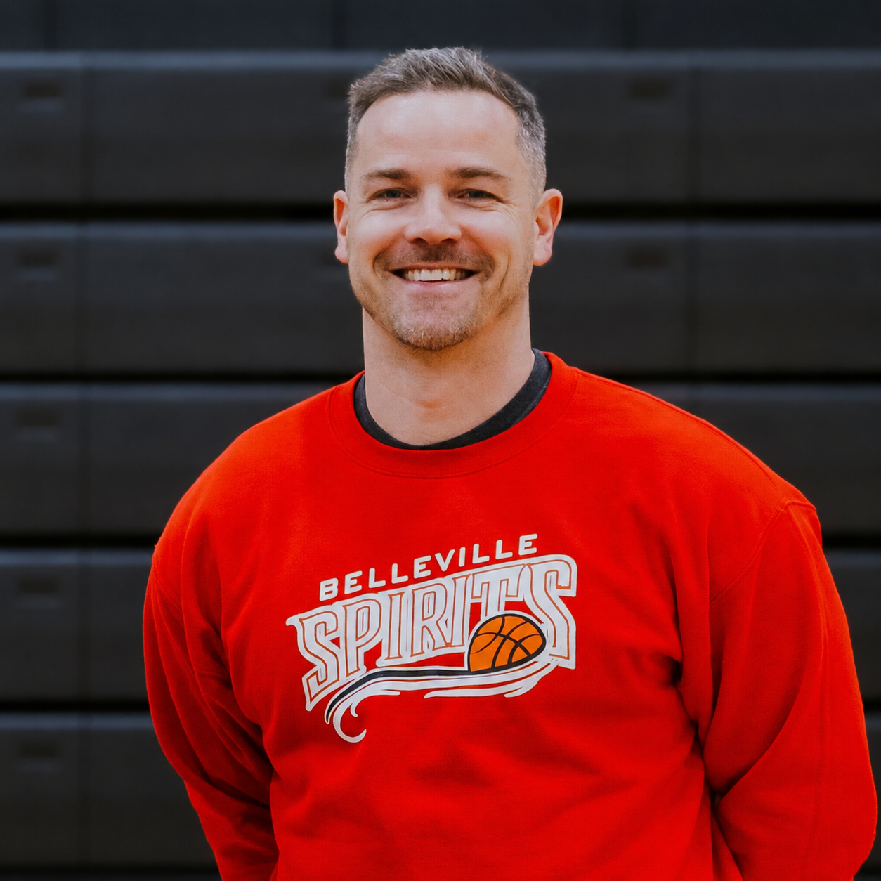 Smiling man in a red Belleville Spirits sweatshirt standing indoors against a dark background