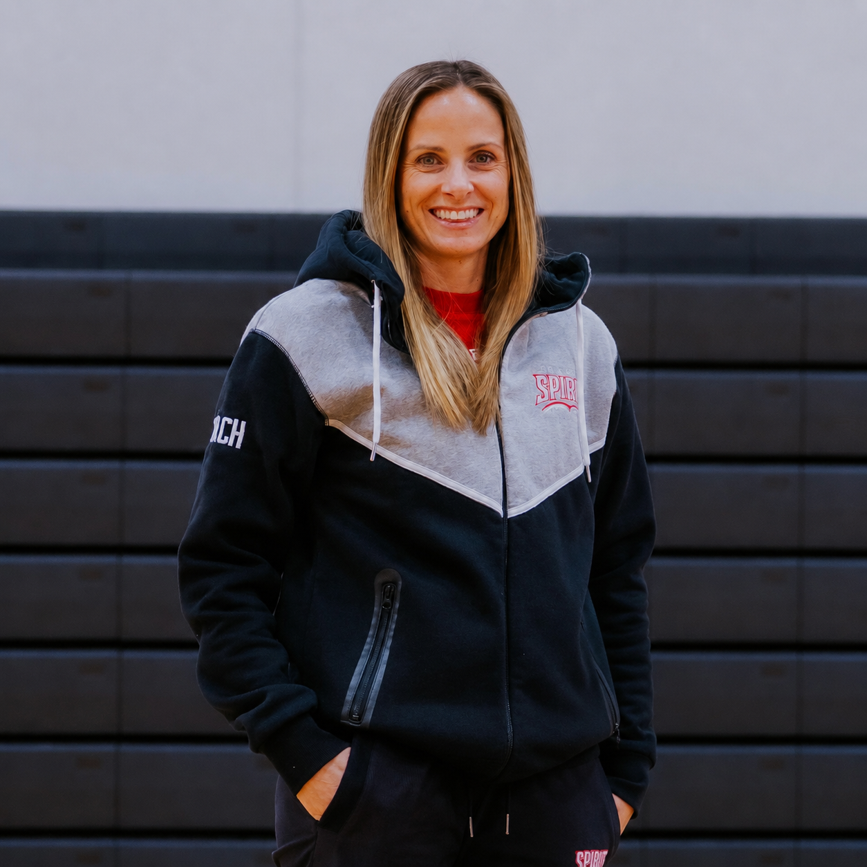 Woman in a black-and-gray hoodie standing in front of a dark wall, smiling at the camera.