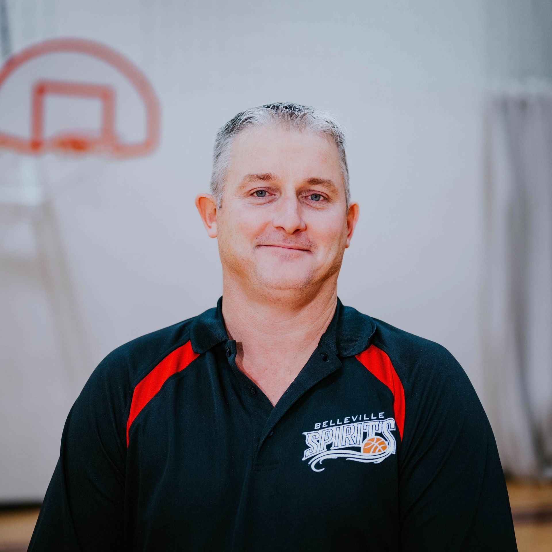 Man standing indoors in a black and red sports shirt, with a basketball hoop blurred in the background
