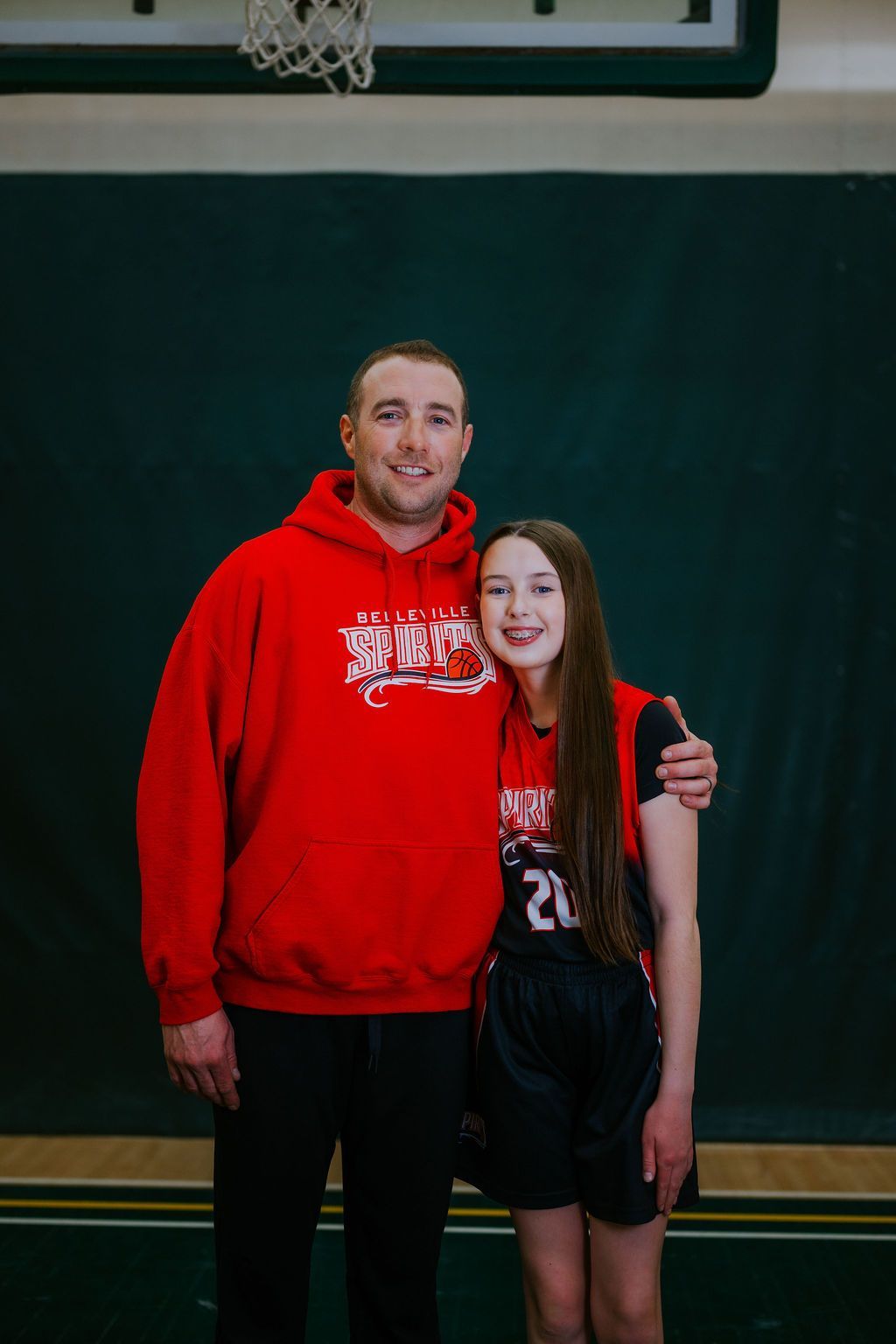 Two people posing in a gym, one in a red hoodie and one in a black basketball uniform, smiling with an arm around each other.