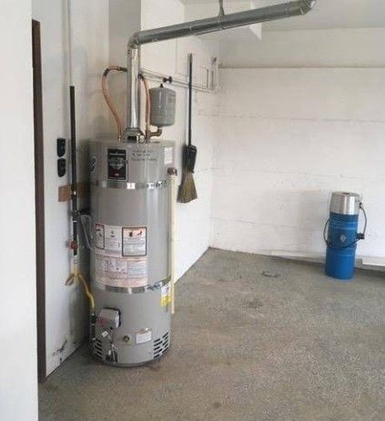 Water heater and pipes in a garage, with a blue barrel in the background.