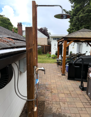 Outdoor patio with brick paving, wooden shower post, and grill under a gazebo in a backyard.