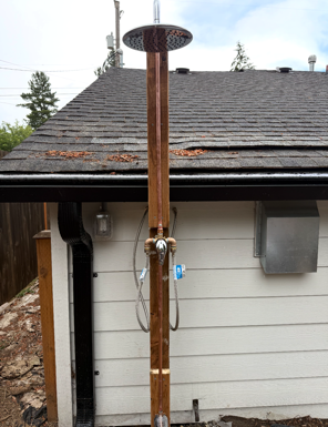 Outdoor shower with rusted metal post and round showerhead beside a house