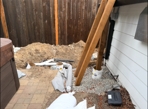 Debris and wooden planks piled in a narrow outdoor corner beside a house and fence