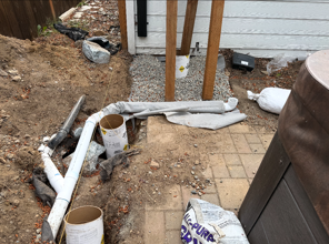 Exposed white drainage pipes and gravel beside a house foundation and patio area.