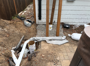 Outdoor construction mess beside a house: white pipes, dirt, and debris near a brick patio and fence