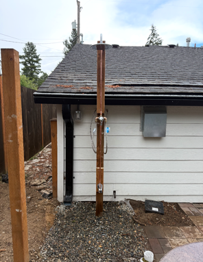 Outdoor wooden shower beside a house, with a hose and gravel ground.