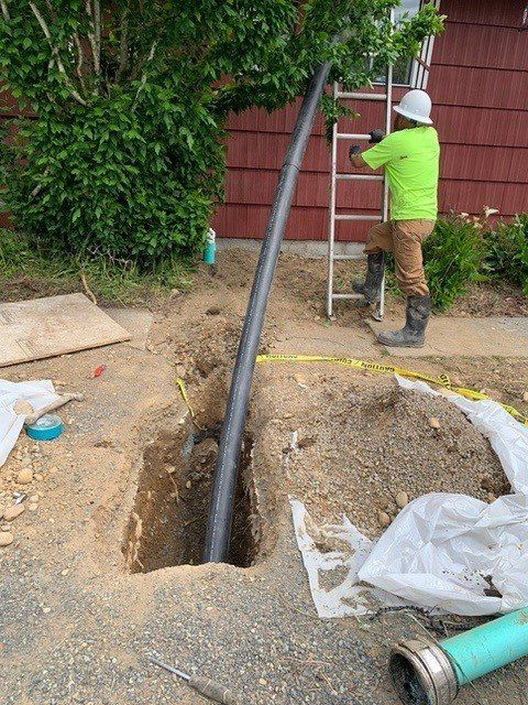 A construction worker in a lime shirt and white hard hat stands on a ladder, installing a large black pipe in a trench.