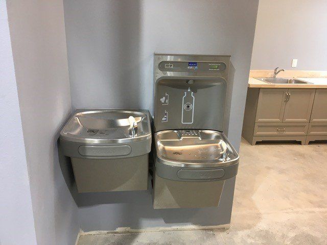 Gray water fountain and water bottle filler unit on a light gray wall with a sink visible in the background.