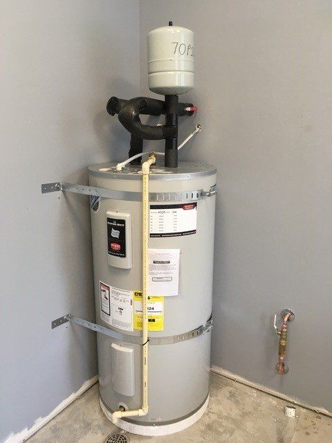 A tall, gray water heater in a corner with a white expansion tank and a bent copper pipe.
