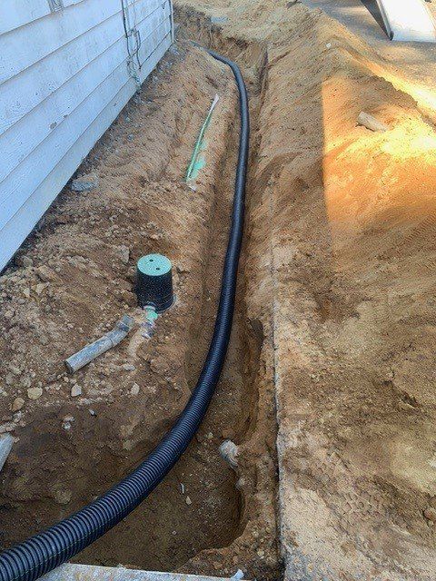A trench alongside a house with black corrugated pipe, a green pipe, and a round green lid.