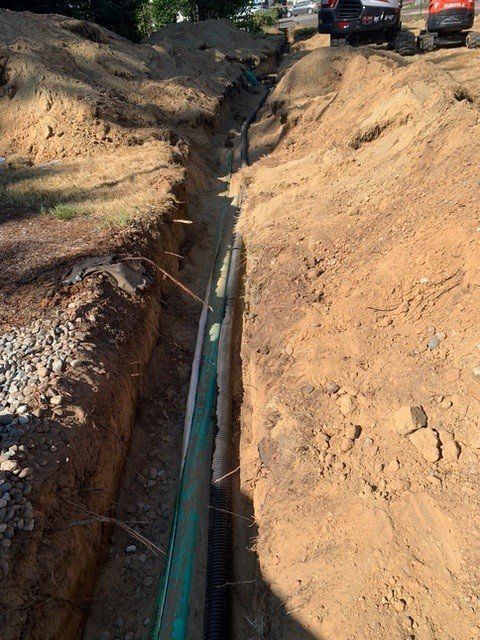 A deep trench dug in the ground, with a green pipe installed. An excavator is visible in the background.