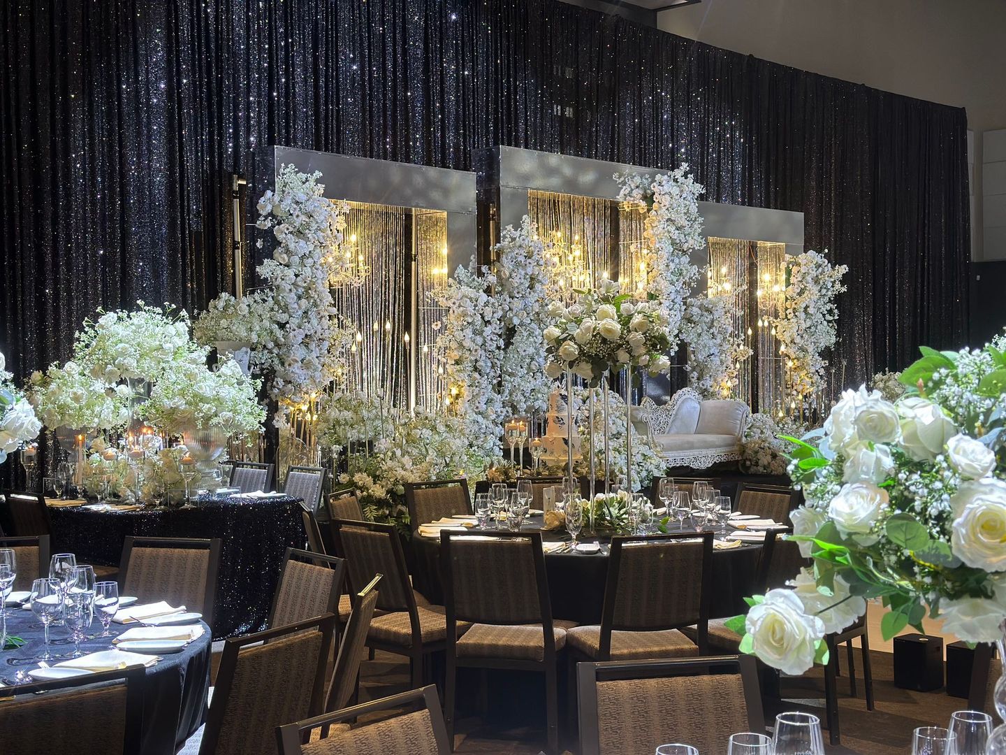 Make your event something you'll never forget! Dark sparkling curtains behind elegant gold, silver, and white displays of flowers and candles on wedding stage before a reception hall.