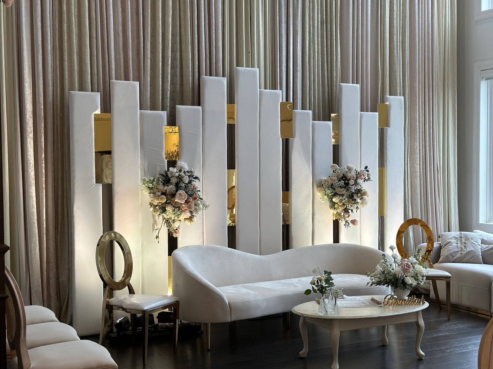 An event stage and backdrop decorated with soft pink curtains and tall standing panels before a divan, chairs, and table