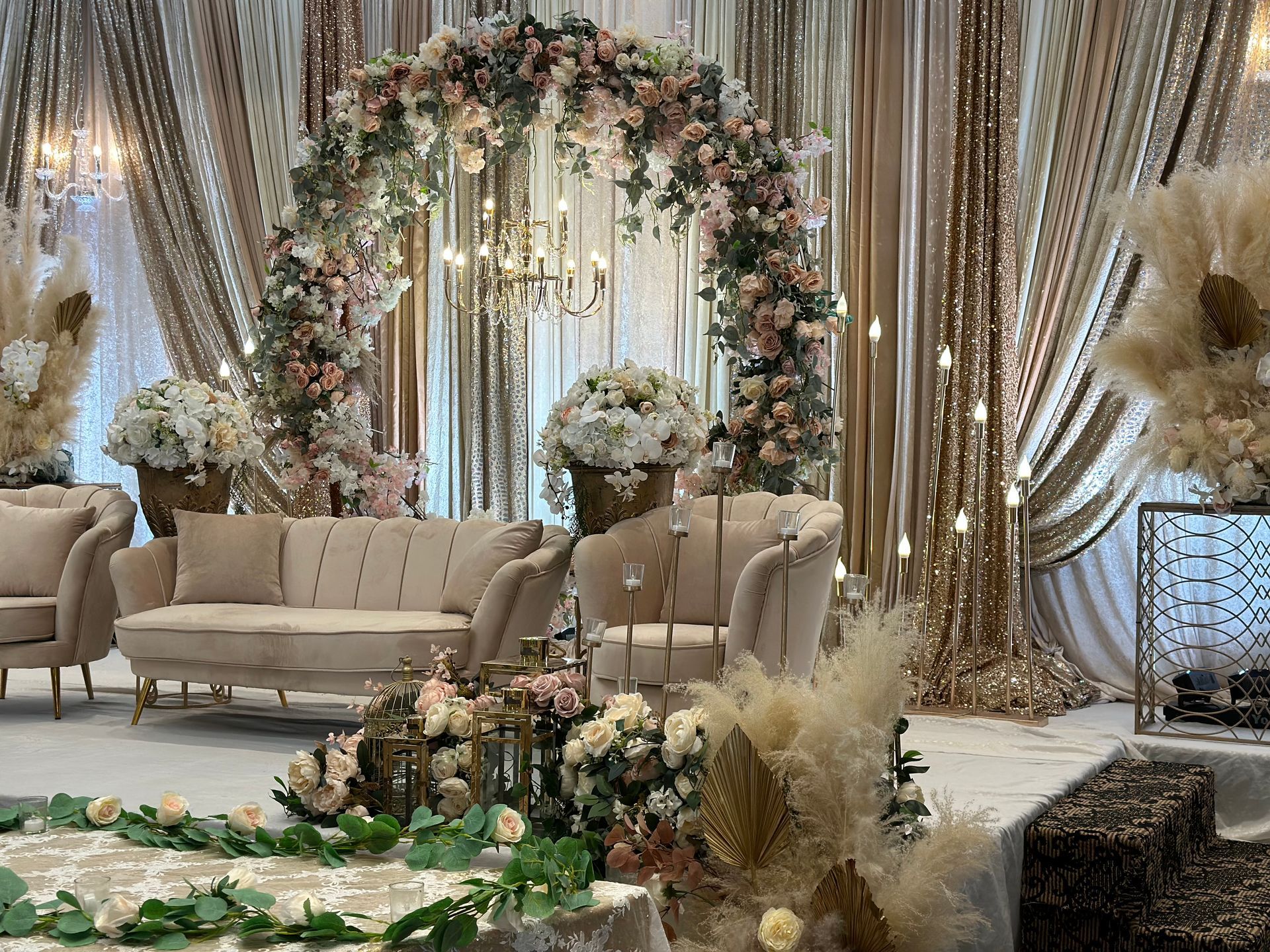 A wedding reception stage with sofas and flowers on it, with a floral canopy above and a drapery backdrop featuring light gold and pink colors