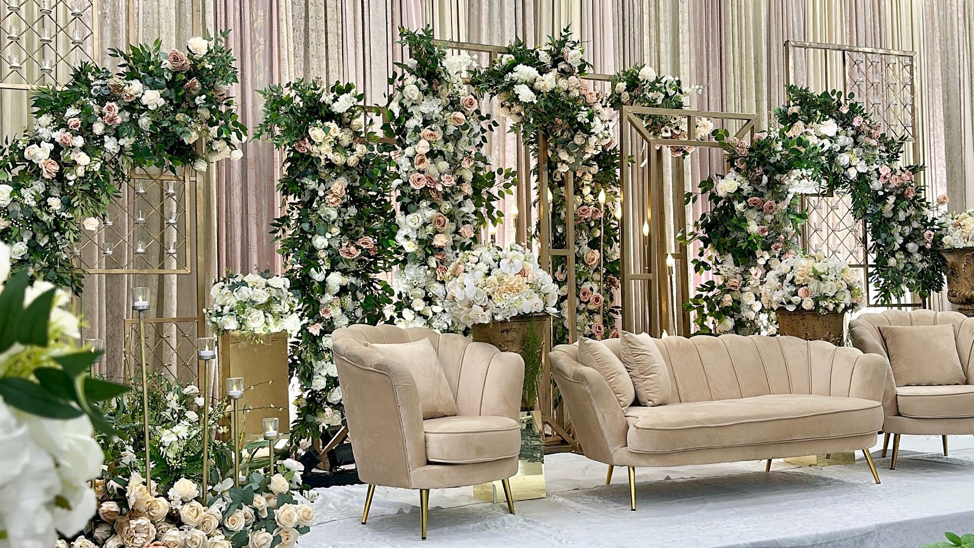 White flowers and vibrant leaves draped around frames above sofas on a reception stage with a luscious curtain in the background