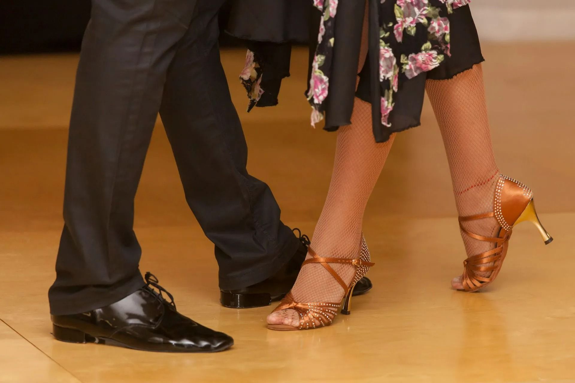 Two dancers’ legs in black and floral attire, stepping on a wooden floor in ballroom shoes.