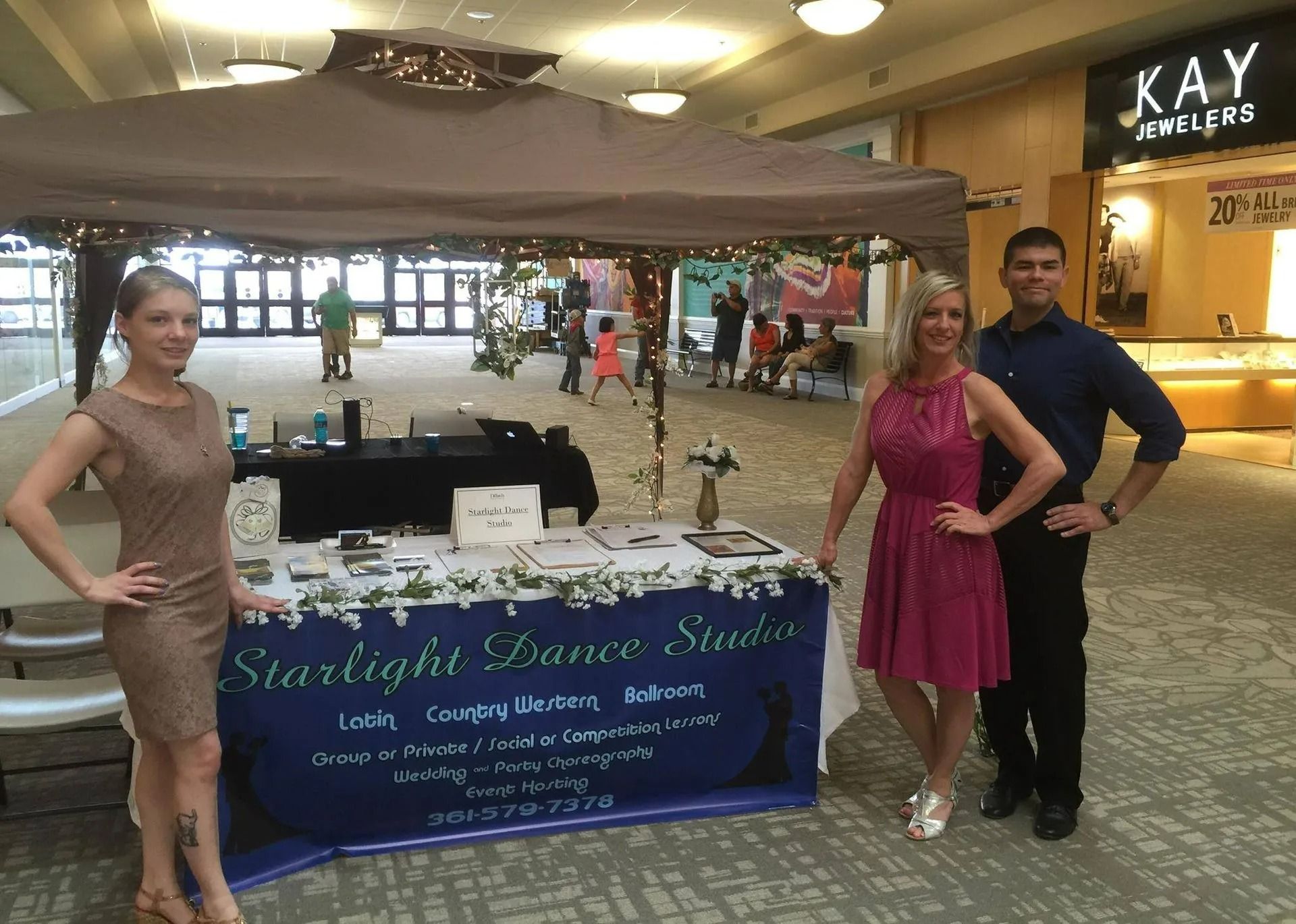 Promotional booth with two women and a man standing beside a table under a canopy in a mall atrium