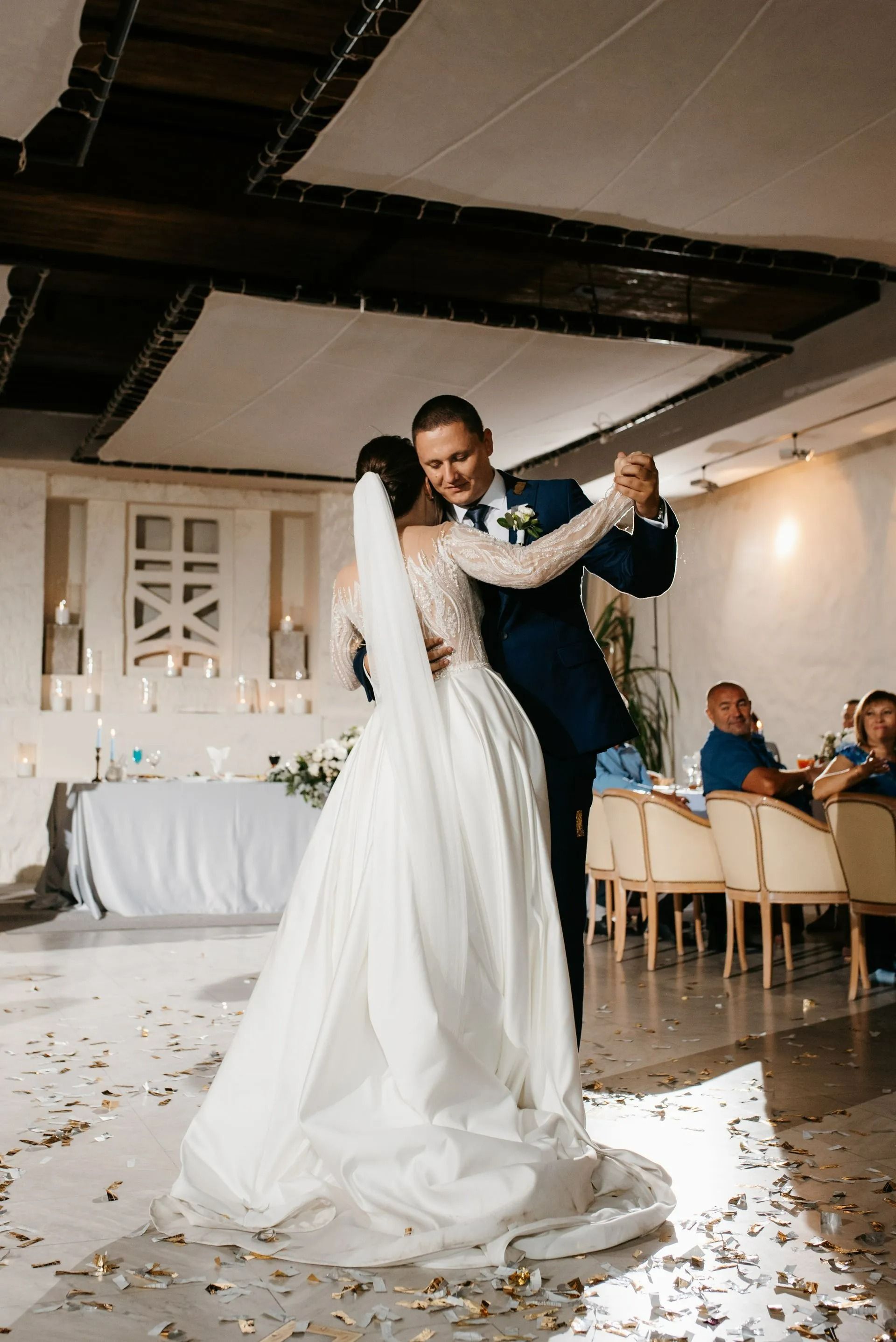 Bride and groom dancing at a wedding reception, with guests seated nearby and confetti on the floor