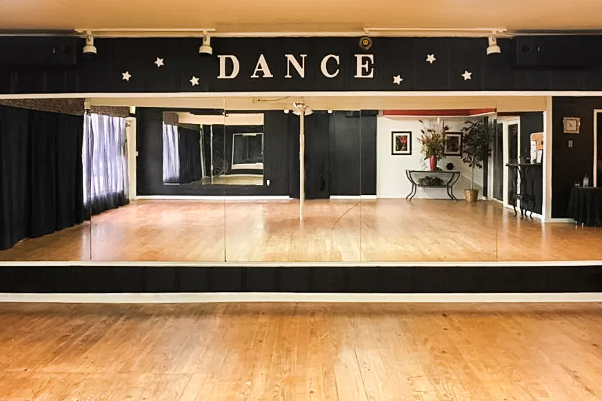 Dance studio with wooden floor, mirrored wall, and black “DANCE” sign above the stage area