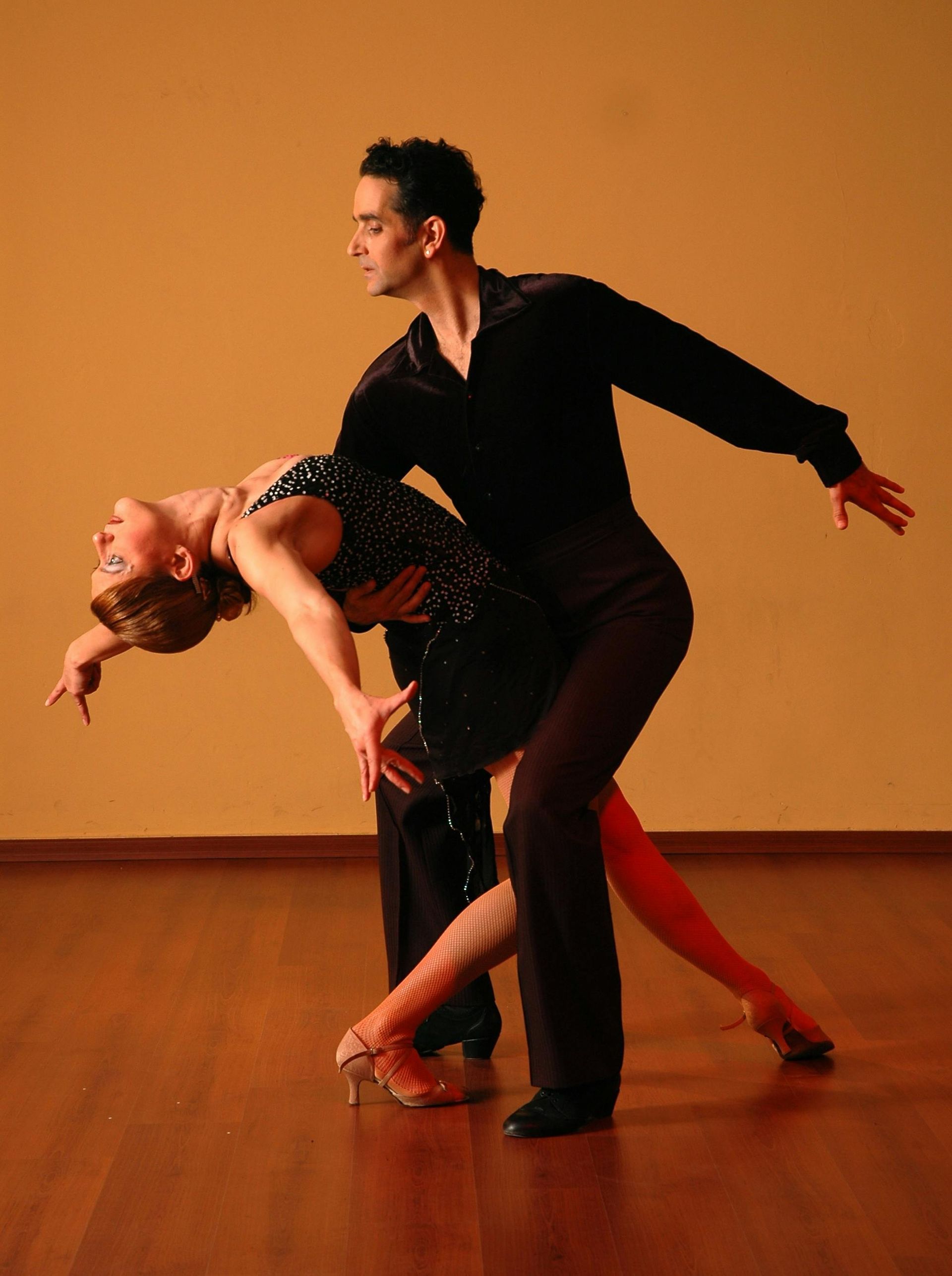 Two ballroom dancers in a dramatic pose on a wooden floor against an orange wall.
