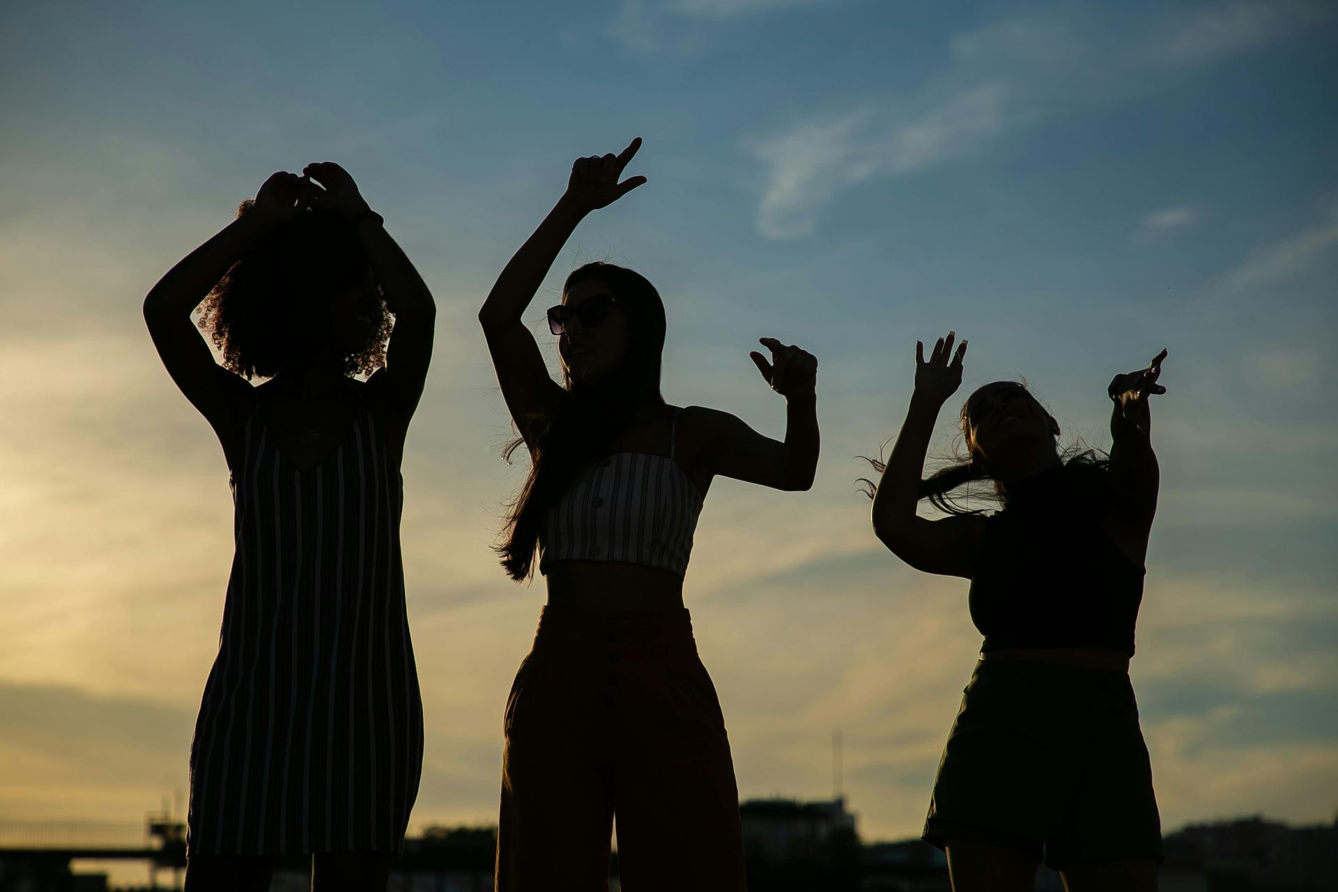 Three silhouetted people dancing with raised arms against a sunset sky