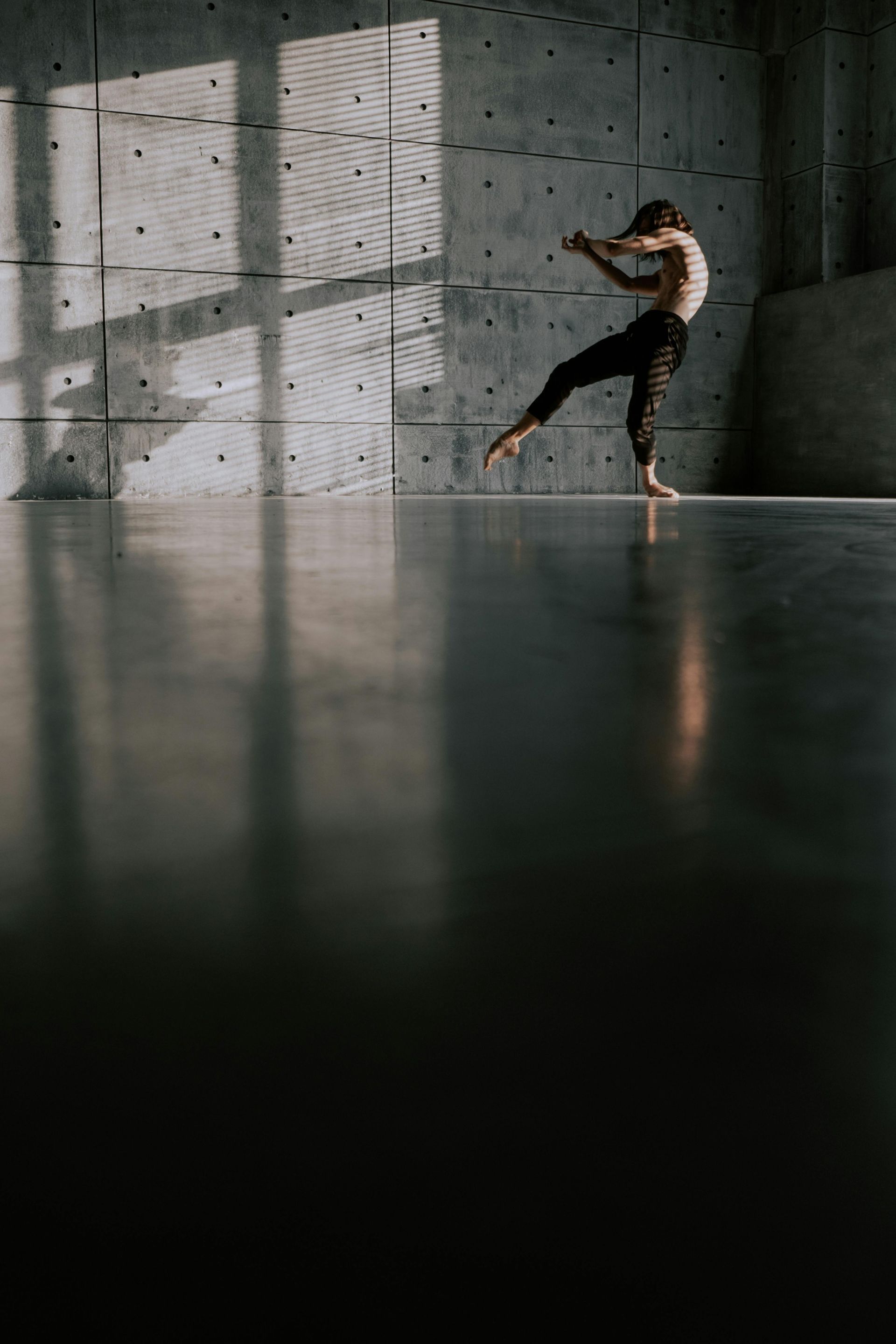 Person dancing in a dim concrete room with tall windows and reflected light on the floor