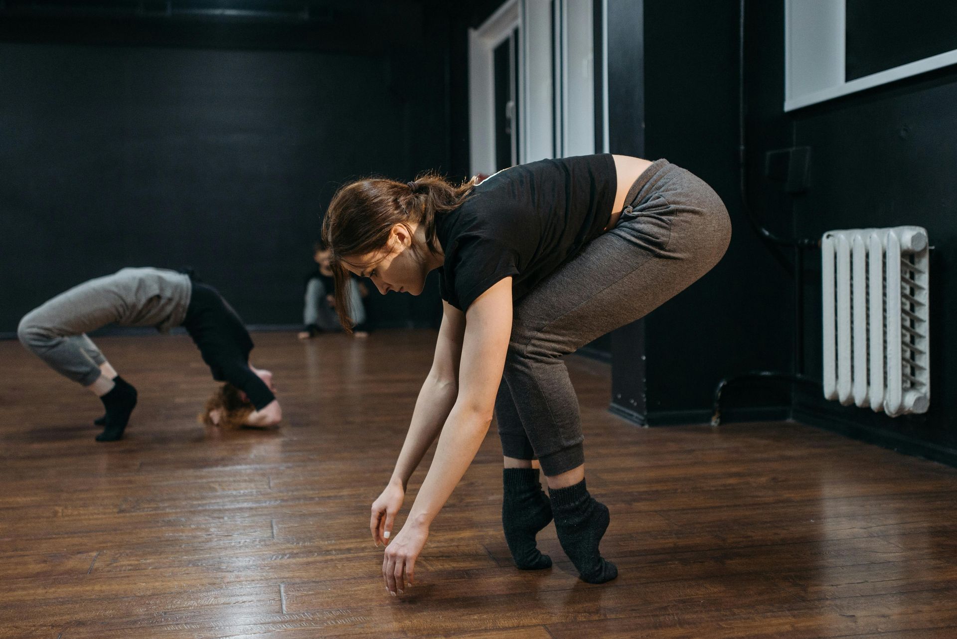 Two dancers stretching on a wooden studio floor, one bent forward with arms reaching down.