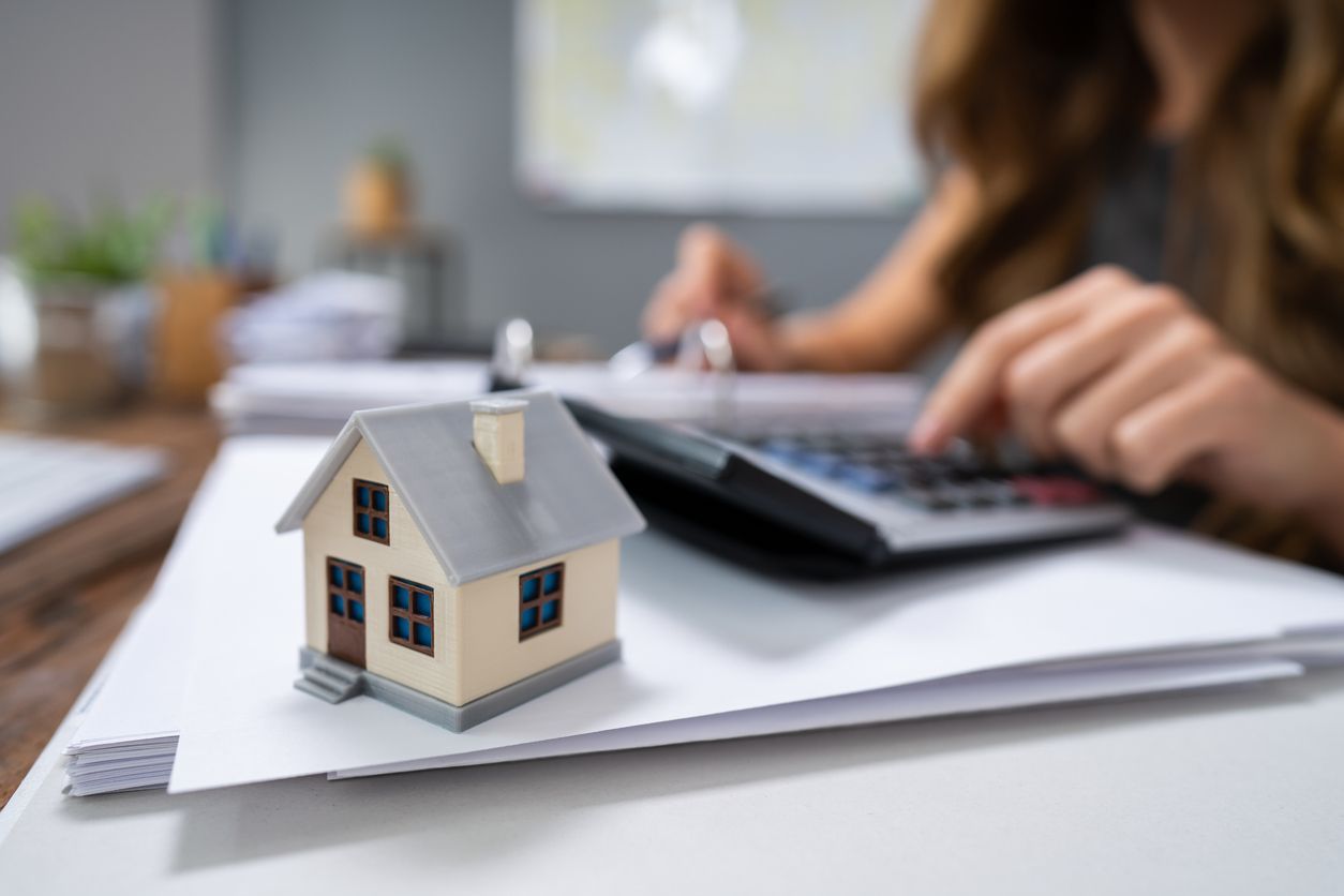 Casa en miniatura sobre papeleo con persona usando una calculadora en el fondo.