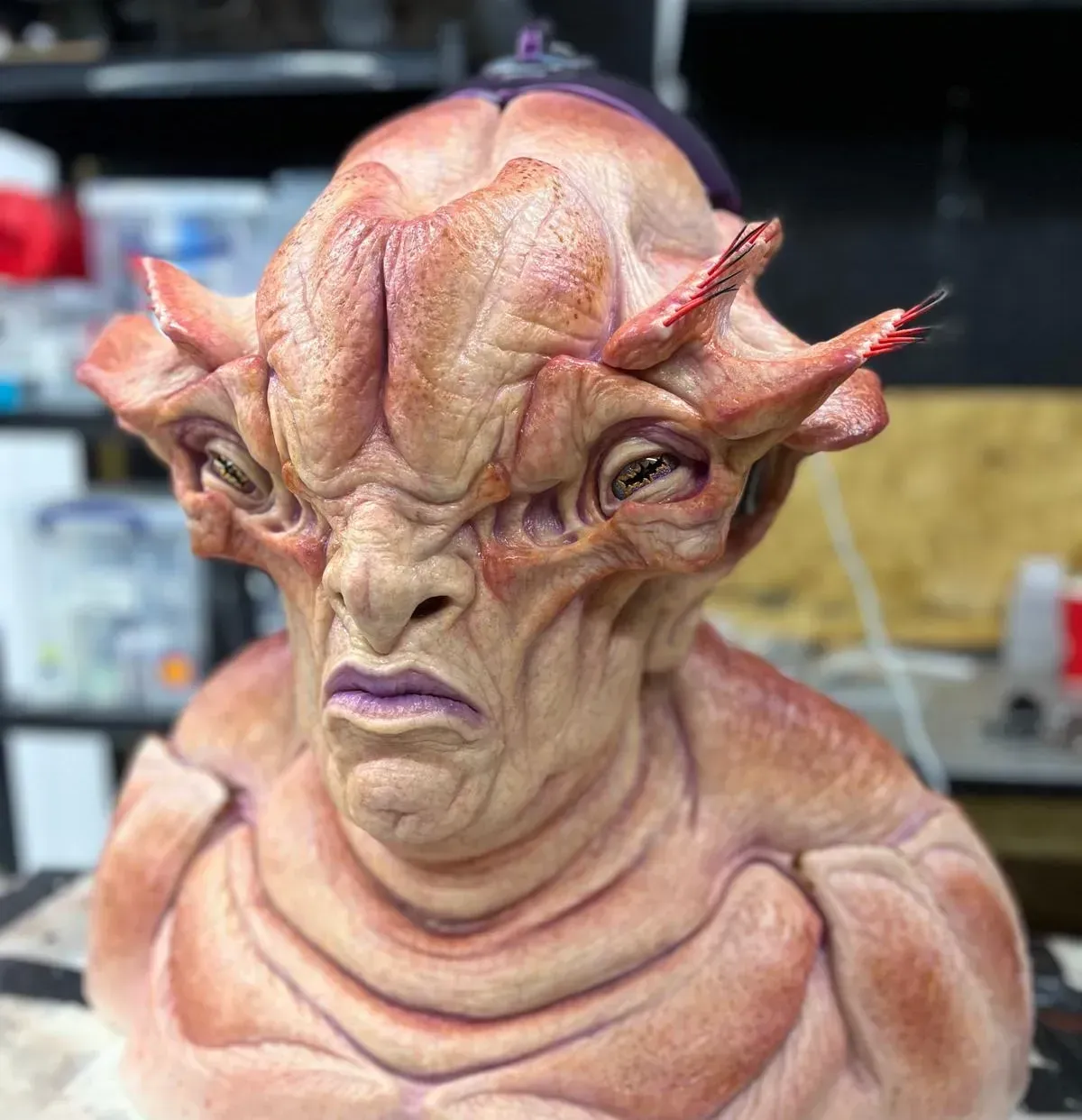 A textured, tan-colored alien bust with prominent head ridges, large eyes, and a somber expression in a workshop setting.