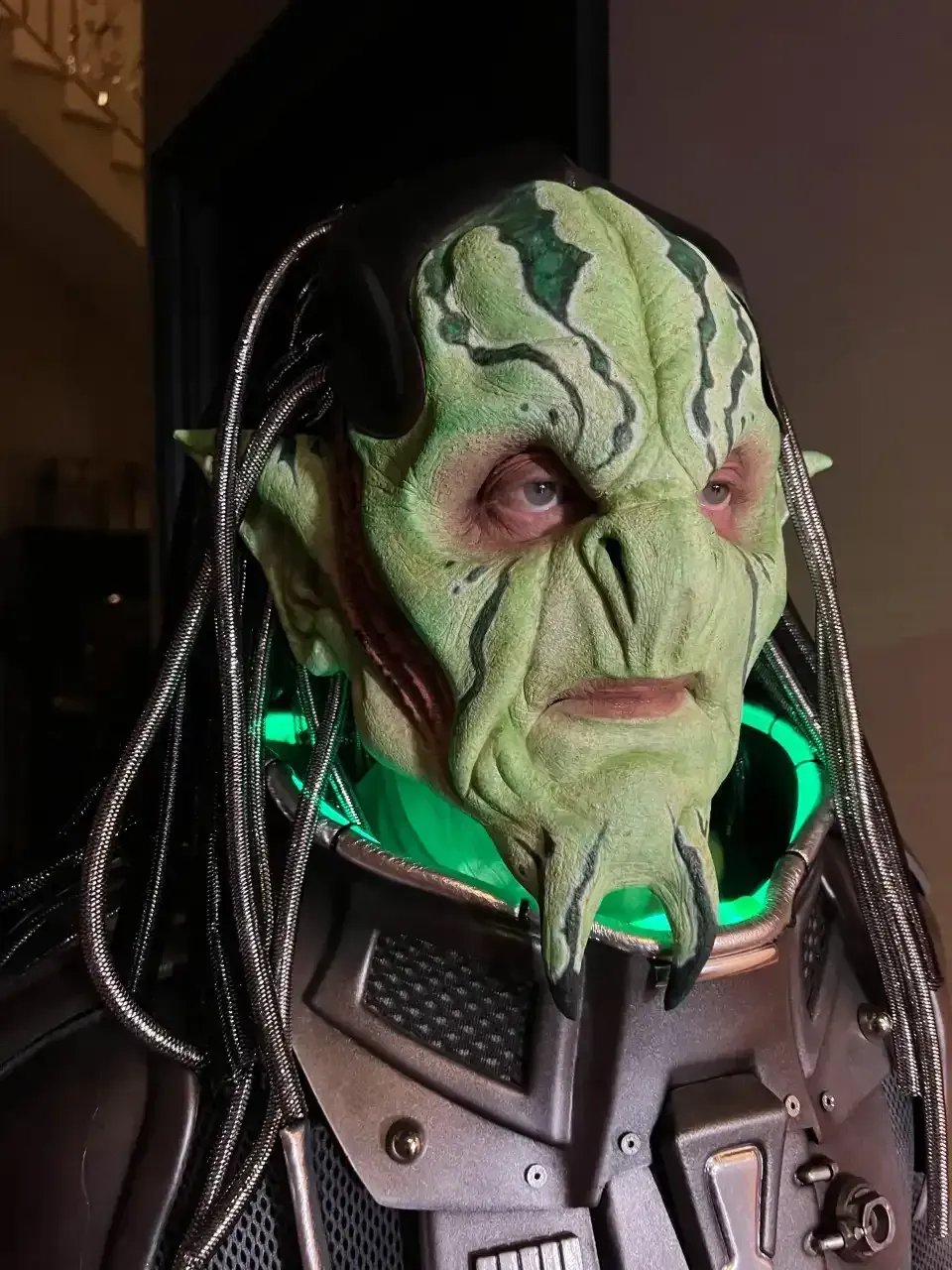 Alien creature with green skin and glowing accents, wearing armor and wires.