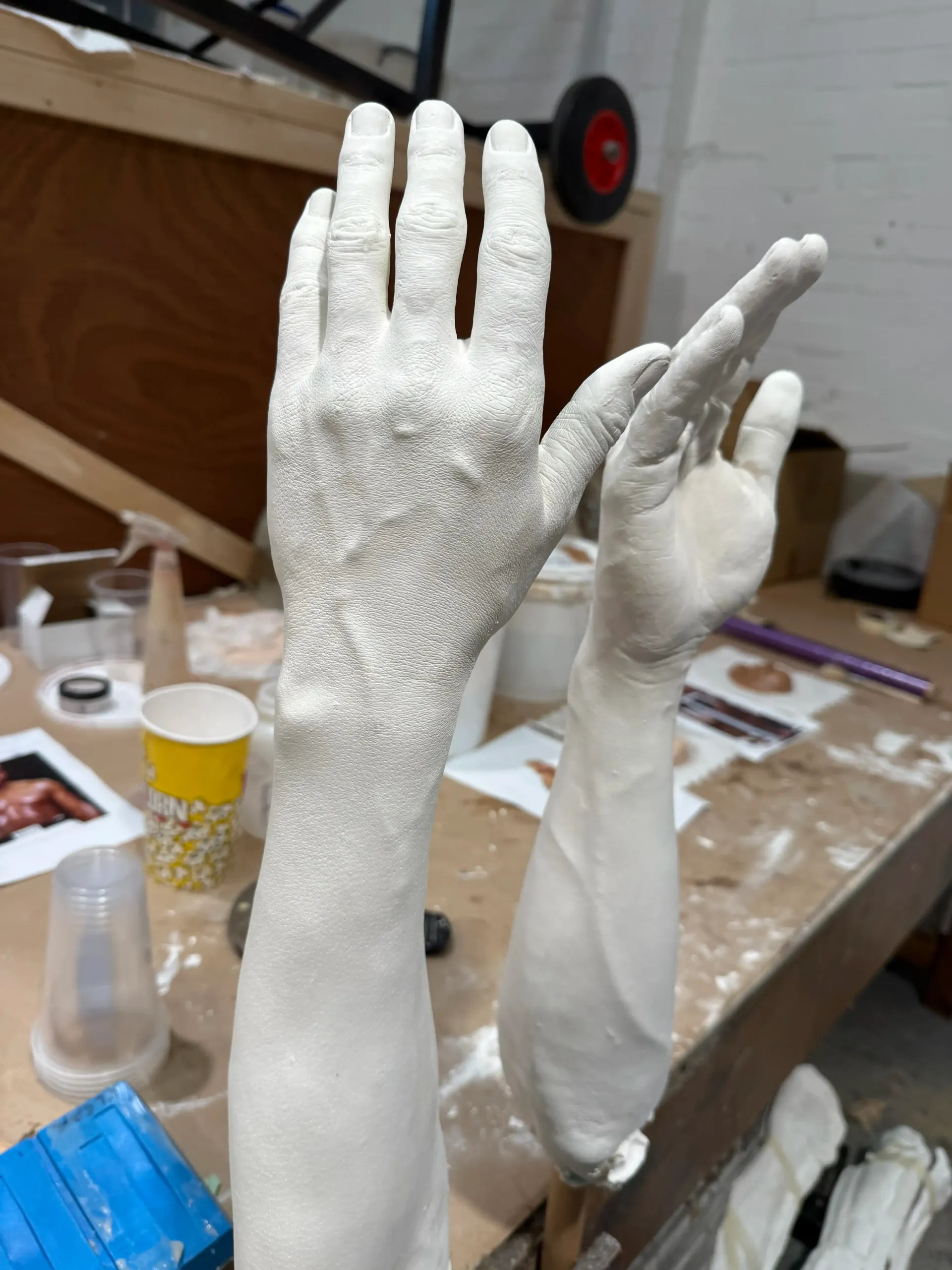 Two plaster-cast sculptures of human arms and hands stand on a cluttered workshop table.