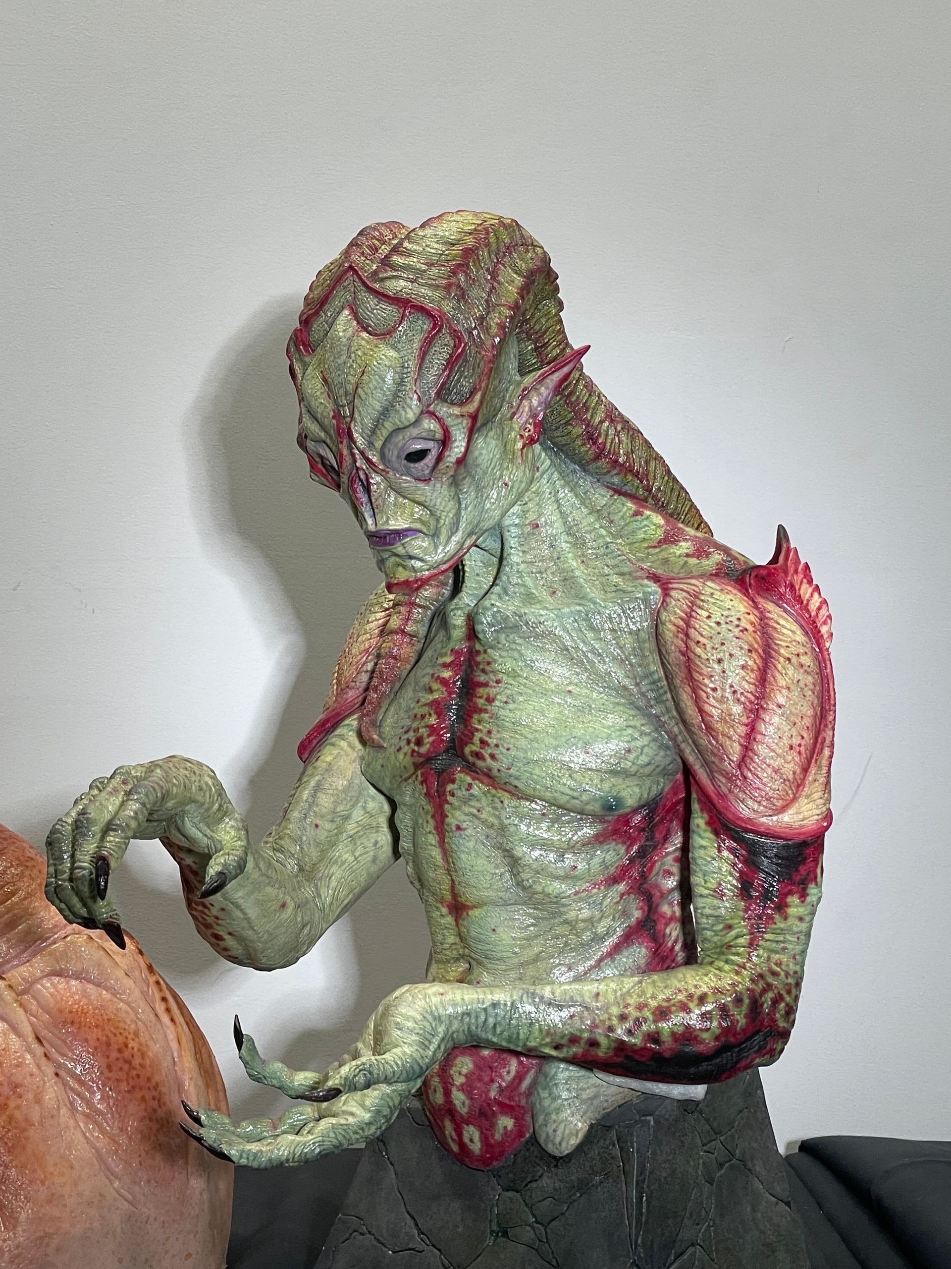A textured, pale green, alien-like bust with red details, tentacles near its mouth, and pointed ears, looking downward.