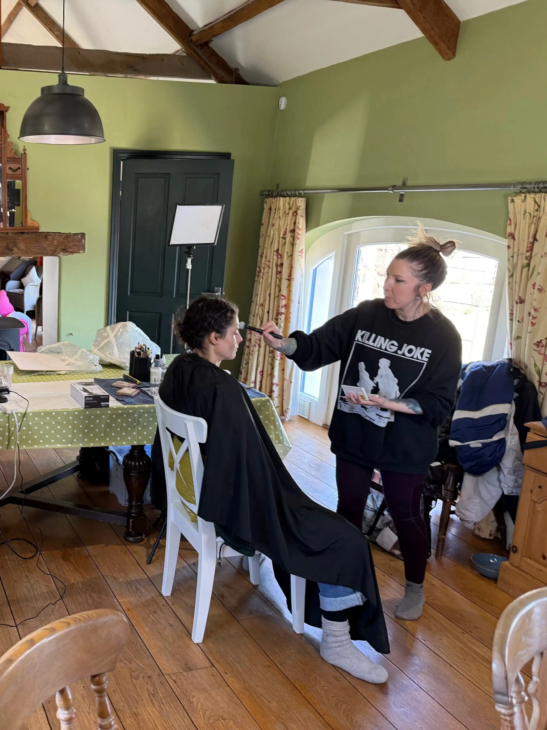 A makeup artist applies makeup to a person sitting in a chair, in a room with a window and a table.
