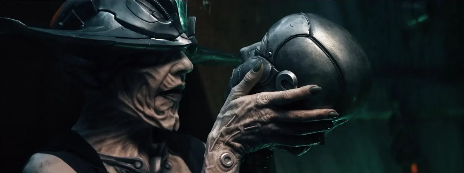 An alien-like figure with wrinkled skin and dark attire holds a metallic, skull-shaped object in a dimly lit, green room.