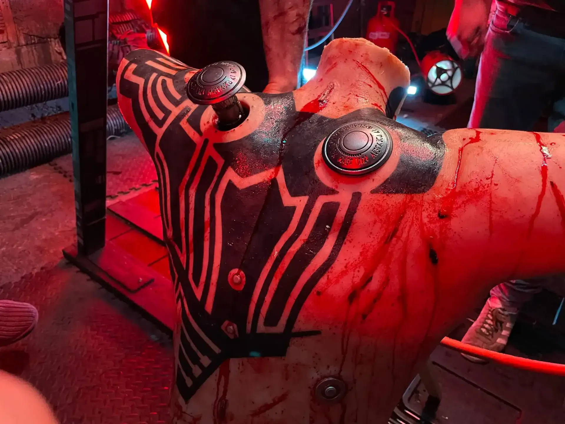 A blood-stained torso mannequin features geometric black markings and metallic circular ports embedded in the skin.