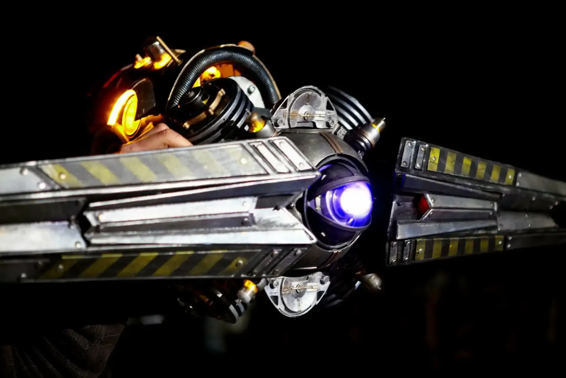 A person holding a sci-fi prop weapon with a glowing purple core and yellow hazard stripes against a black background.