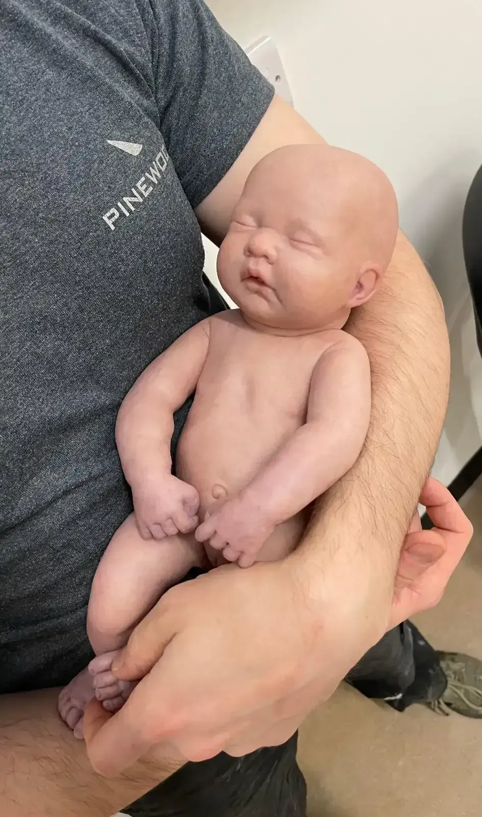 A person in a gray t-shirt holding a small, realistic doll of a newborn baby in their arms.