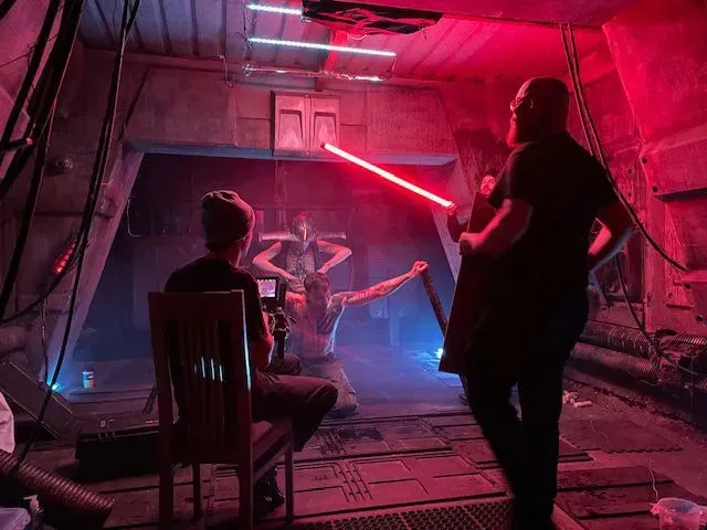 Behind-the-scenes film set with a person holding a red light stick, a camera operator on a chair, and a person posing.