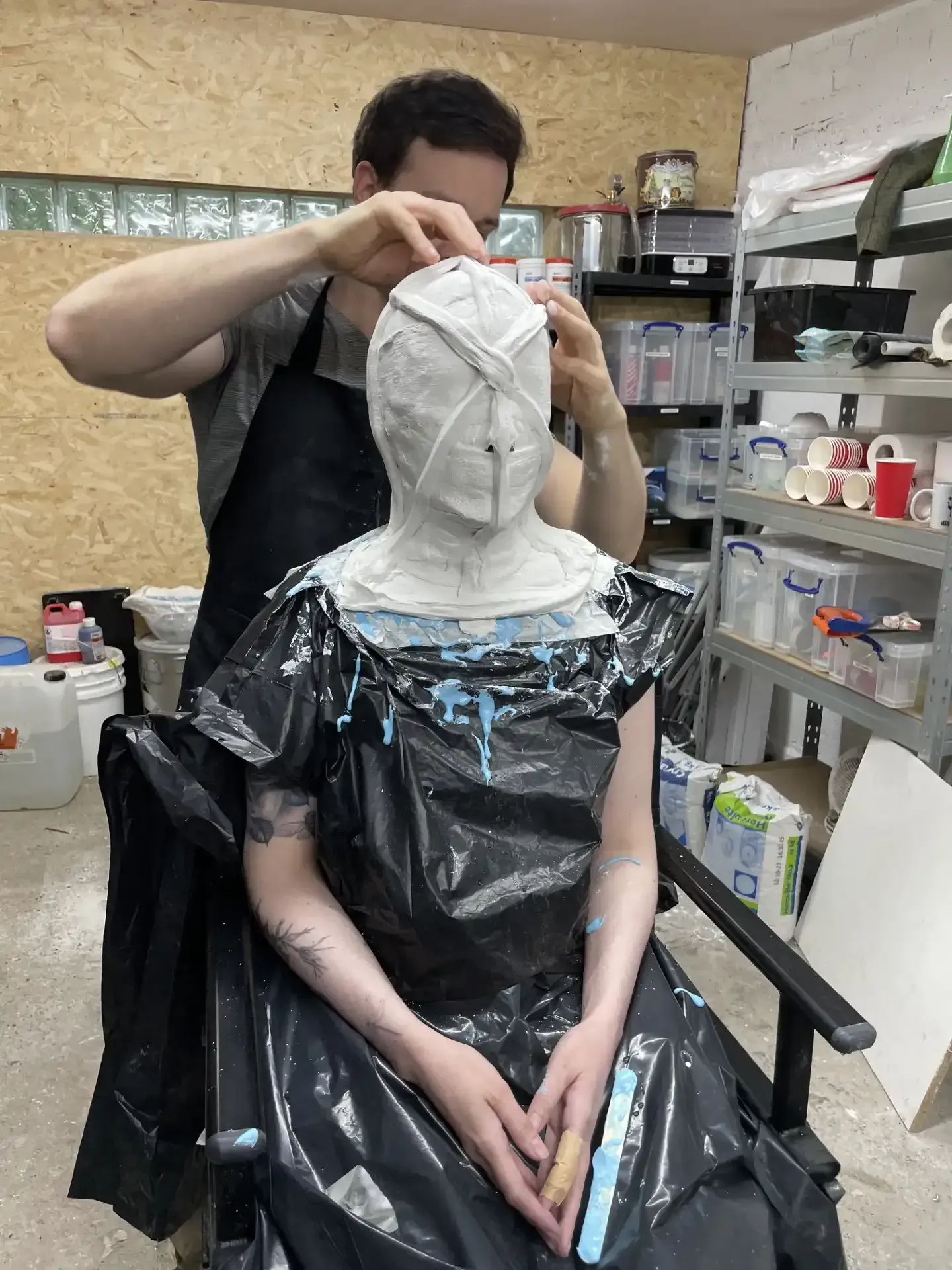A person applies wet plaster bandages to a seated person’s head, both wearing black aprons in a workshop setting.