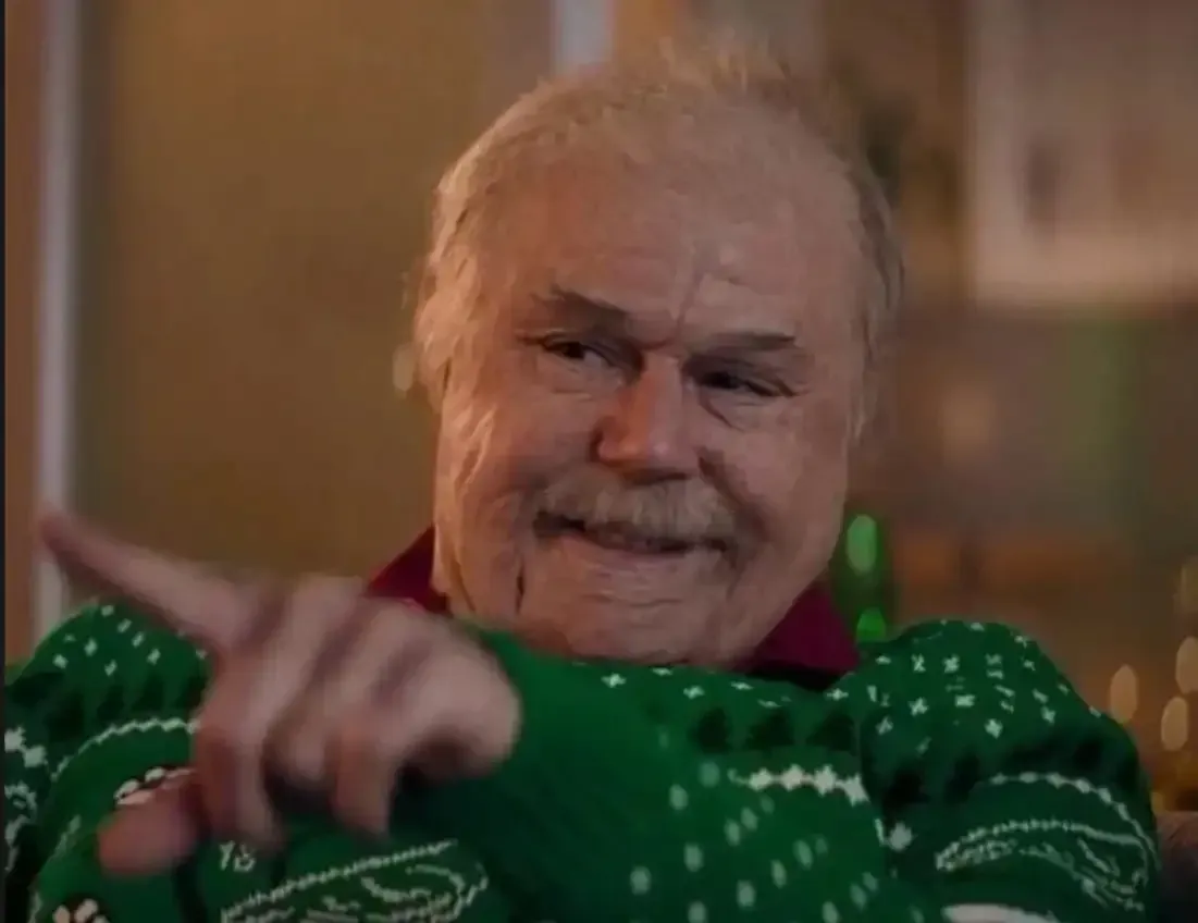 An older person with a mustache, wearing a green patterned sweater, points to the side with a smiling expression.